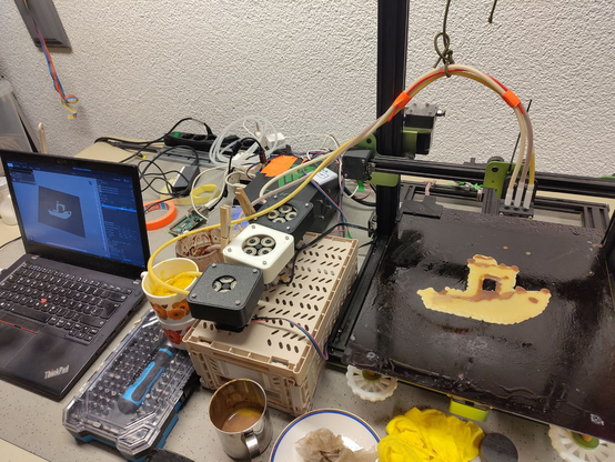 Der Pfannkuchendrucker mit einem 2D-Benchy-Pfannkuchen in weiß mit braunen Akzenten auf dem Druckbett. Daneben stehen Gläser mit weißem, braunem und gelbem Teig. Schläuche verbinden die Gläser mit dem Druckkopf. Neben den Gläsern steht ein Laptop, auf dem PrusaSlicer geöffnet ist und das Benchy-Modell anzeigt