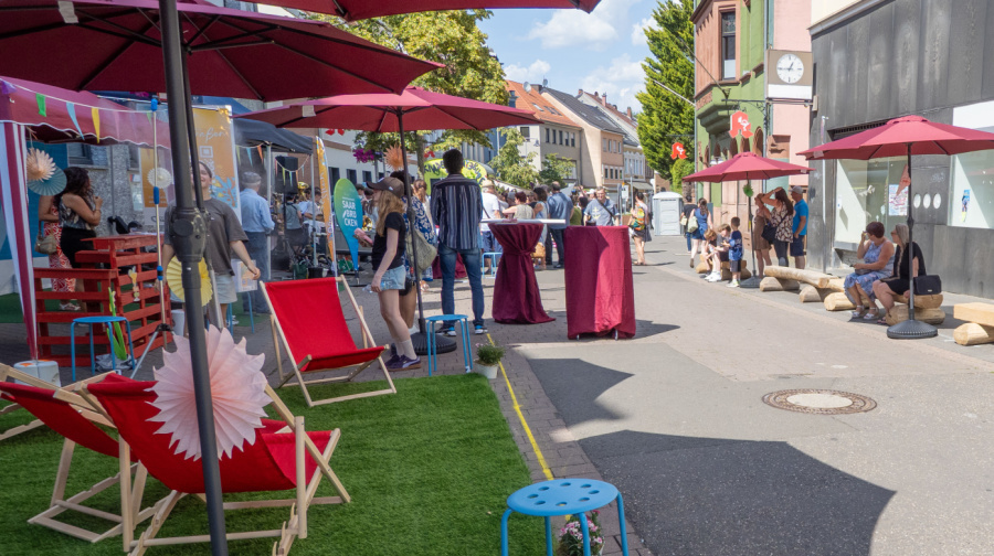 Ein Foto der belebten Straße, mit Sitzgelegenheiten, Zelten, einer Grünfläche, Sonnenschirmen und Menschen.